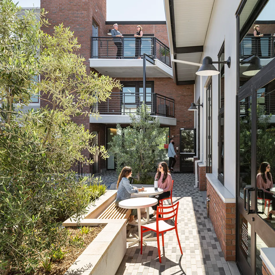 Two women sitting and talking at a round table with red chairs in a sunny outdoor courtyard surrounded by brick and white buildings, while two people chat on a balcony above.