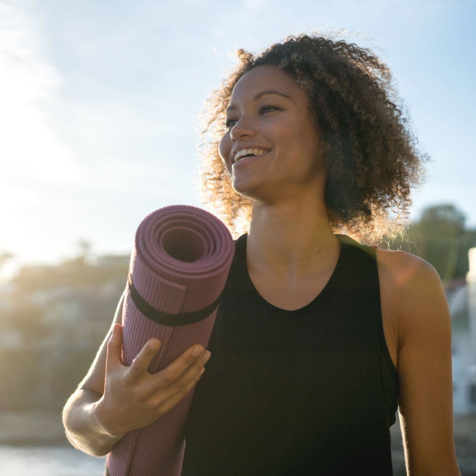Smiling woman holding a rolled-up yoga mat outdoors at sunrise or sunset.
