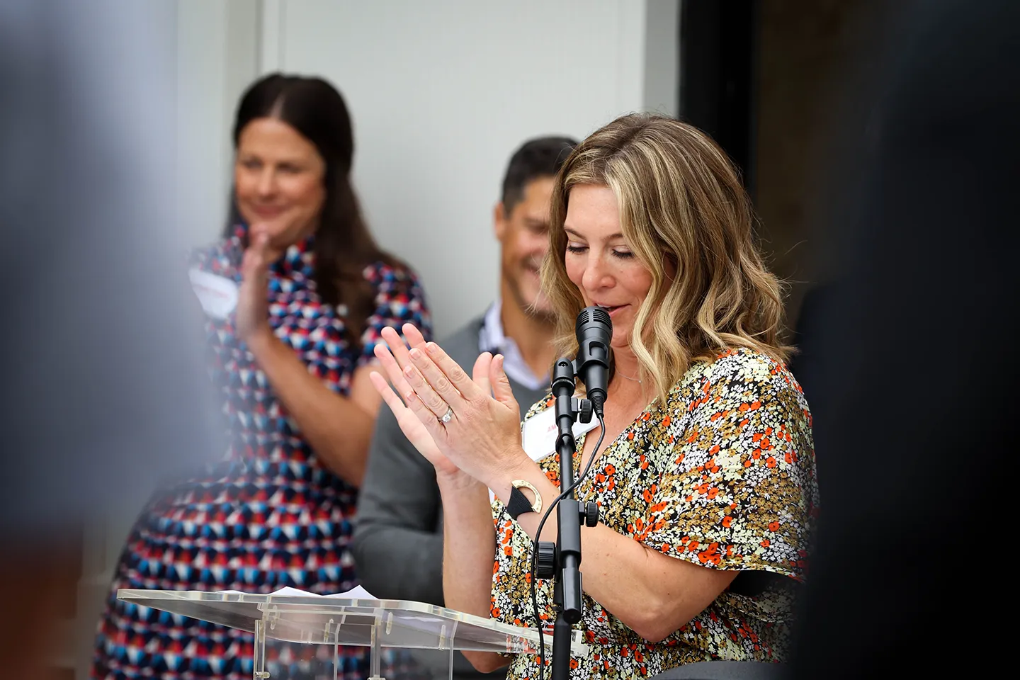Woman with wavy hair in a floral dress clapping and speaking into a microphone at a podium with two people clapping in the background.