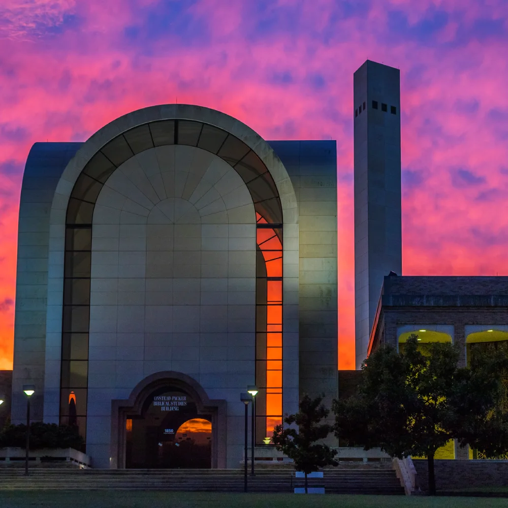 Sunset-lit biblical studies building with large arched window and adjacent tower under a purple and orange sky.