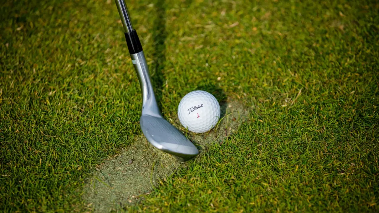 Close-up of a Titleist number 1 golf ball on grass next to a wedge club head positioned in a divot on the turf.