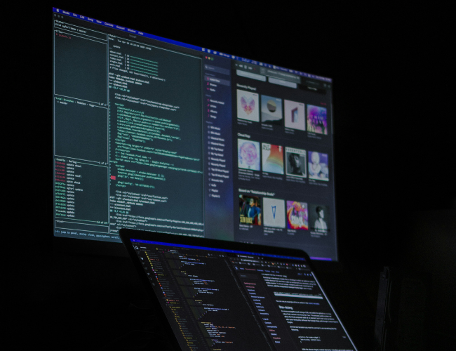 Two monitors in a dark room displaying lines of code and an image gallery, with a laptop open below the main screen.