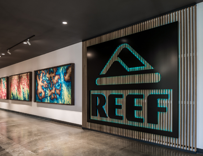 A modern lobby featuring a large illuminated REEF logo on a slatted wood wall, alongside framed colorful coral-inspired artwork.
