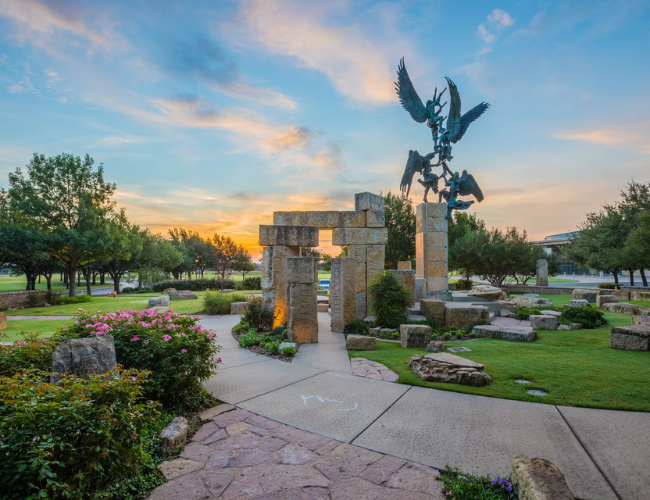 Outdoor university plaza at sunset with a bronze eagle sculpture and stone archway surrounded by landscaped trees.