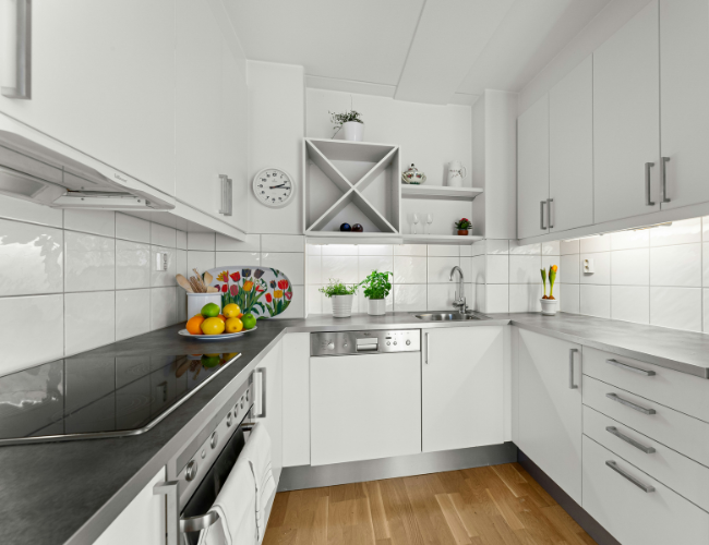 Bright white U-shaped kitchen with gray countertops, white tile backsplash, stainless appliances, and fresh herbs and fruit on the counter.