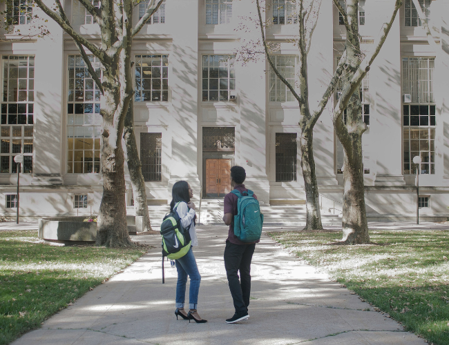 Two students with backpacks stand on a walkway in front of a large classical university building, framed by bare trees on a sunny day.