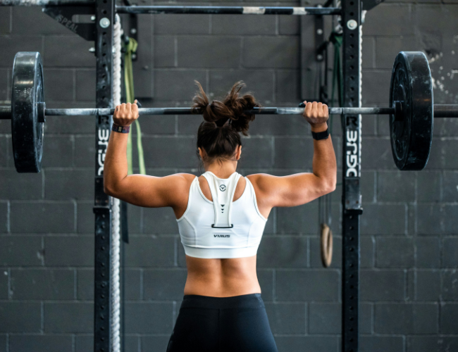  Rear view of a woman in athletic wear lifting a loaded barbell overhead at a squat rack in an industrial-style gym.
