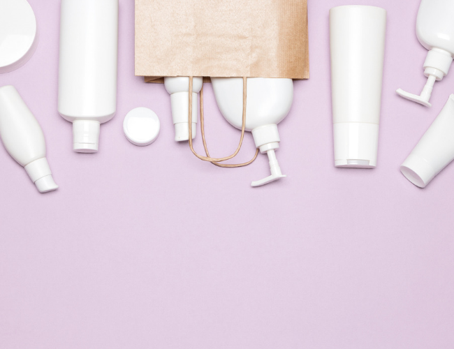 Flat lay of white personal care product bottles arranged around a brown paper shopping bag on a lavender background.