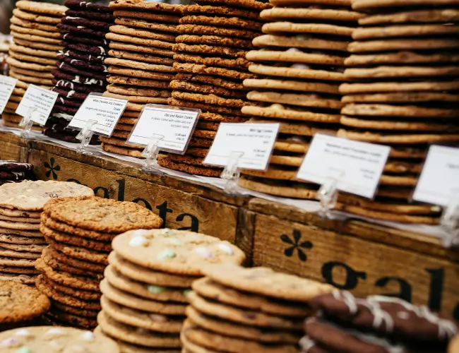Tall stacks of assorted fresh-baked cookies displayed on wooden crates at a bakery, with handwritten flavor labels in front of each variety.
