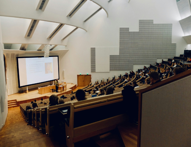 Large lecture hall filled with students watching a presentation on a large screen at the front of the room.