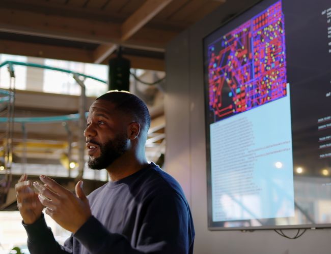 A man gestures while presenting beside a large screen displaying a colorful circuit board diagram and lines of code.
