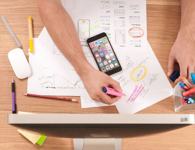 Overhead view of a designer sketching mobile app wireframes on paper with highlighters, surrounded by a smartphone, pens, and a monitor.