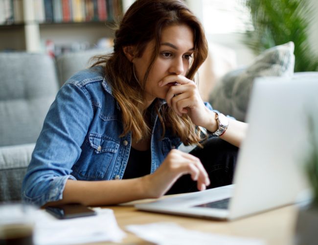 Woman in a denim jacket resting her hand on her chin while thoughtfully reading content on a laptop in a sunlit living room.