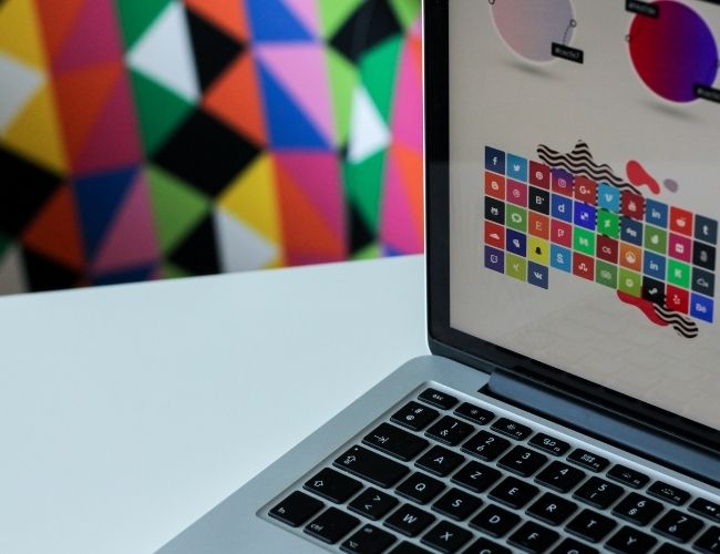 Open MacBook displaying a mosaic of social media app icons next to a colorful geometric triangle wall art on a white desk.