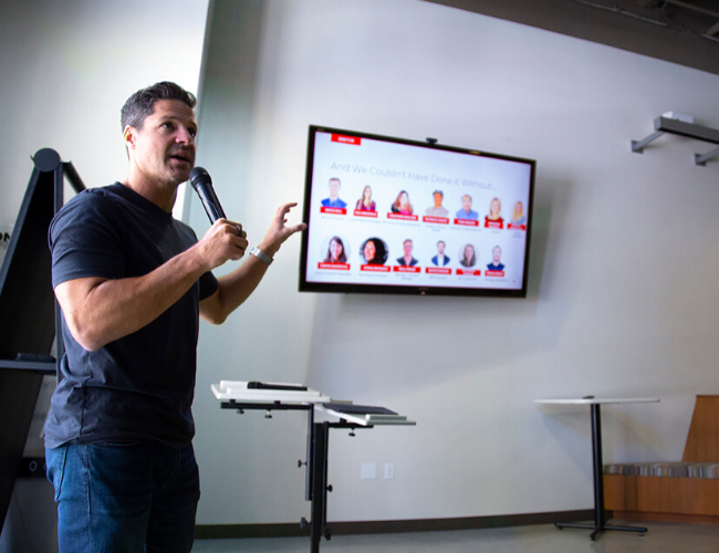 Reid Carr speaking at the front of a conference room holding a microphone, with a TV screen showing a panel of people behind him.