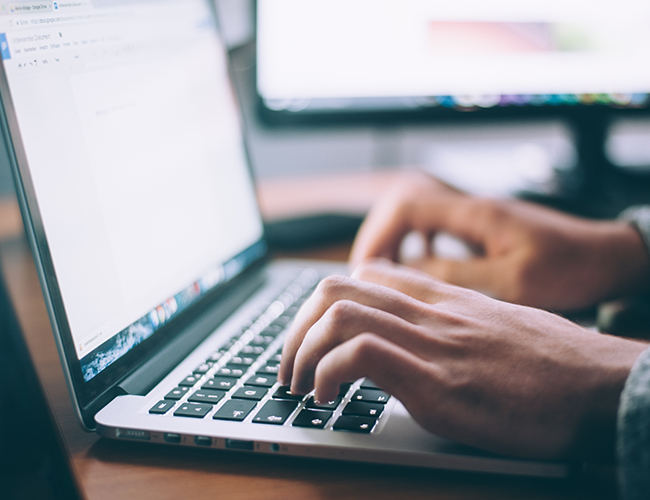 Hands typing on a MacBook laptop keyboard with a second monitor visible in the background.