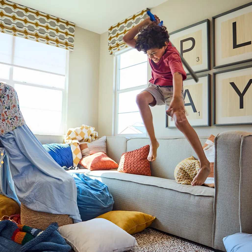 Child joyfully jumps on a couch with a toy sword in a colorful, pillow-filled room, embodying playful adventure and imagination