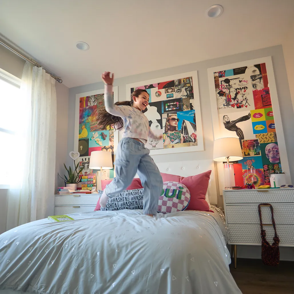 Girl joyfully jumps on bed in colorful room with vibrant wall art, pink pillows, and a bright window, capturing playful energy.