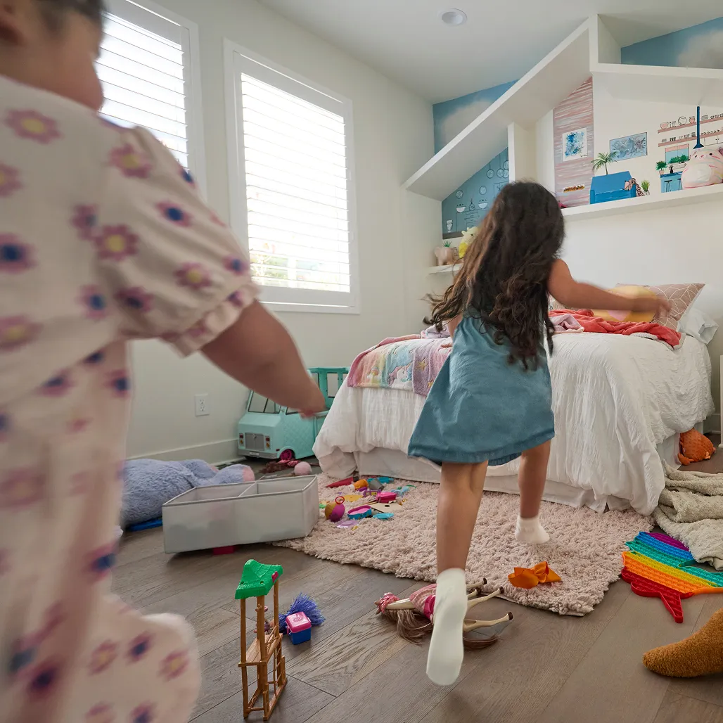 Two children play energetically in a colorful, toy-filled bedroom with a cozy bed and creative wall decor
