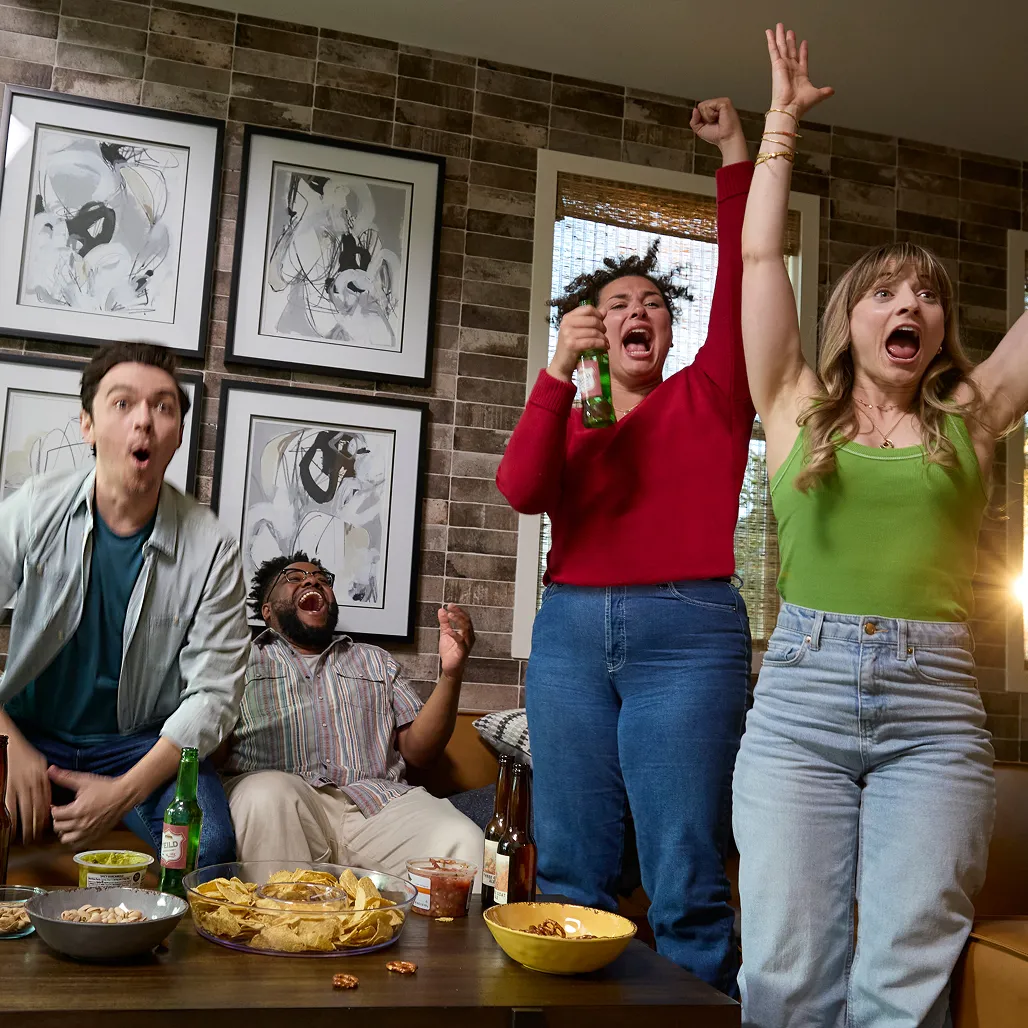 Four friends cheer energetically on a couch, surrounded by snacks and drinks, celebrating a sports victory in a cozy living room.