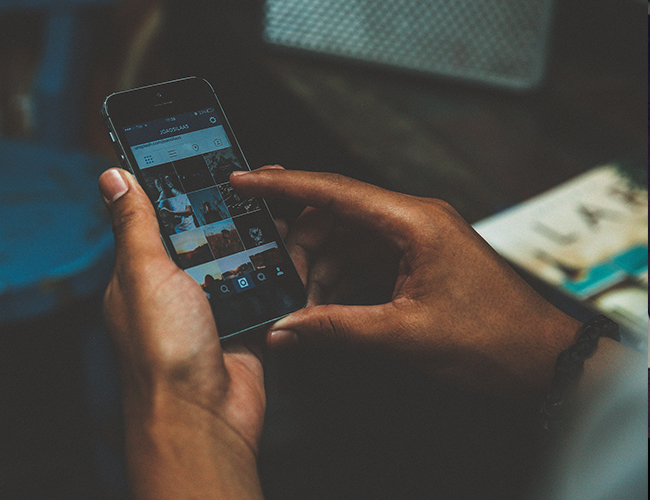 Hands holding a smartphone scrolling through an Instagram photo grid in a dimly lit setting.