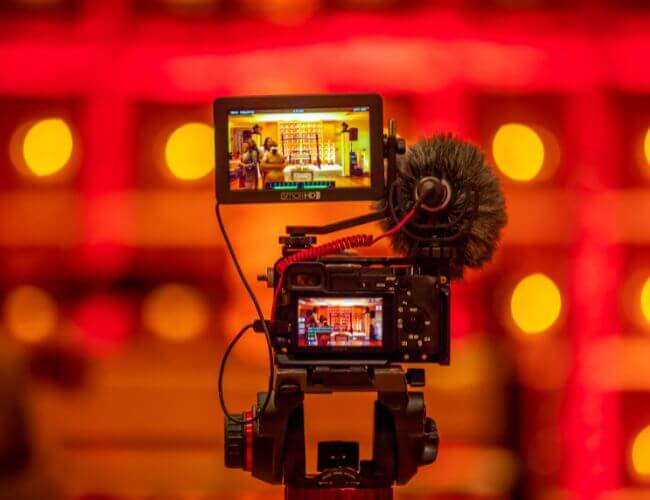 Video camera mounted on a tripod with an external monitor and shotgun mic filming a brightly lit red event with bokeh background.