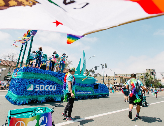 Colorful parade float with rainbow flags and SDCCU branding moving down a sunny street lined with a crowd.