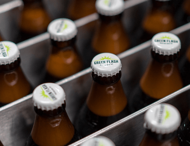 Rows of Green Flash Brewing Co. beer bottles with branded white caps on a stainless steel production line.