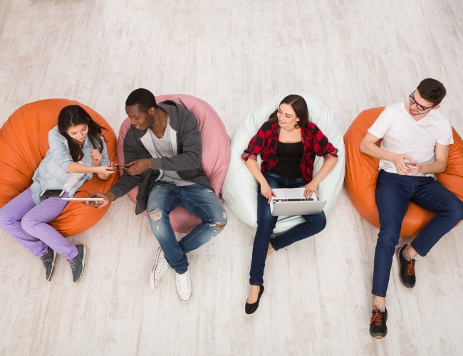 Overhead view of four diverse young people sitting on colorful bean bag chairs, each using a laptop, tablet, or smartphone.