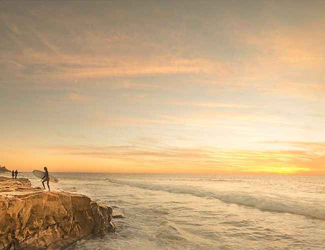 Two figures standing on a rocky coastal cliff overlooking the ocean at sunset with golden sky and waves.
