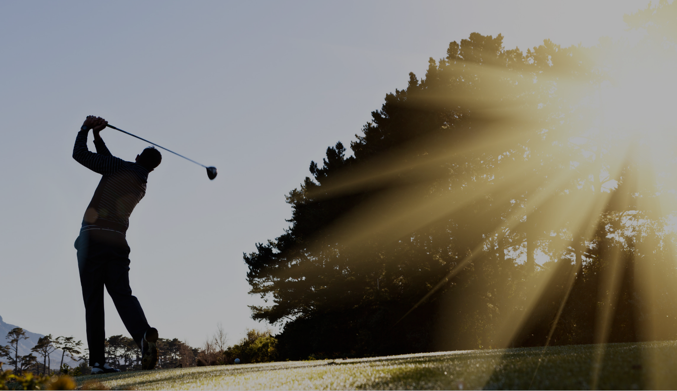Silhouette of a golfer mid-swing with sunlight streaming through trees in the background.