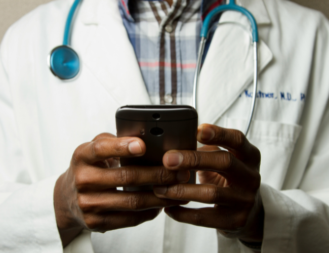 Close-up of a doctor in a white coat with a blue stethoscope around his neck, using a smartphone with both hands.