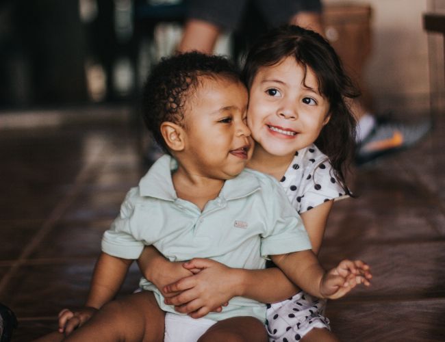 Two young children hugging while seated on a tiled floor, both smiling.