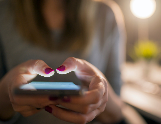 Close-up of a woman with red nail polish texting on a smartphone in warm low light.