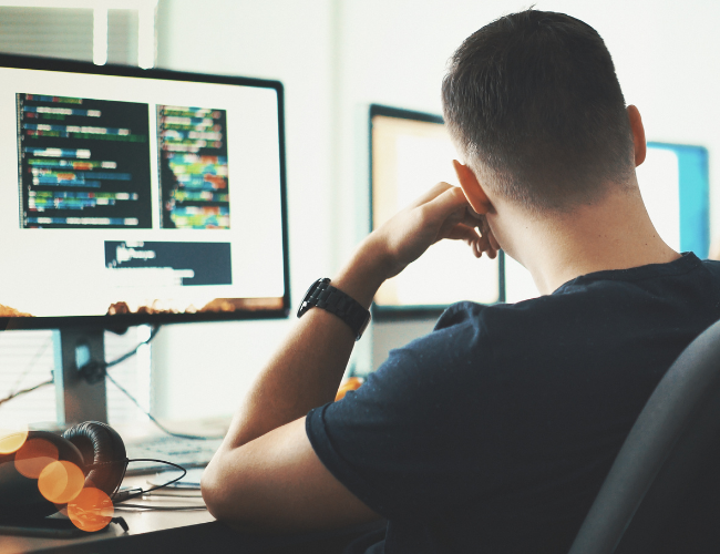 Young person from behind sitting at a desk looking at dual monitors displaying lines of code in a dim room.