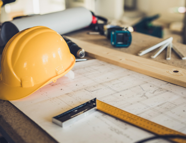 Yellow construction hard hat resting on architectural blueprints with a ruler, pencil, and wood plank on a desk.