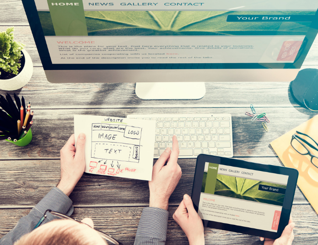 Overhead view of hands using a tablet displaying a website mockup on a wooden desk with glasses, plants, and a printed page.