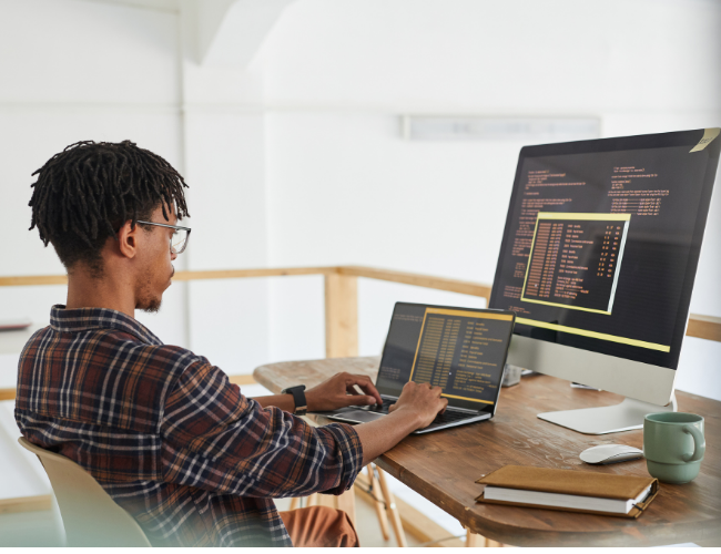 Young man with dreadlocks coding on a laptop while a large monitor in front of him displays lines of code in a bright room.