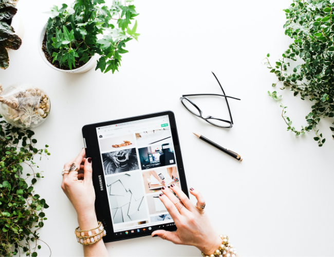 Hands browsing a content grid on a tablet surrounded by potted plants, glasses, and a pen on a white surface.