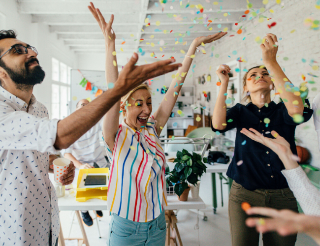 Group of diverse coworkers laughing and celebrating with confetti falling in a bright office.