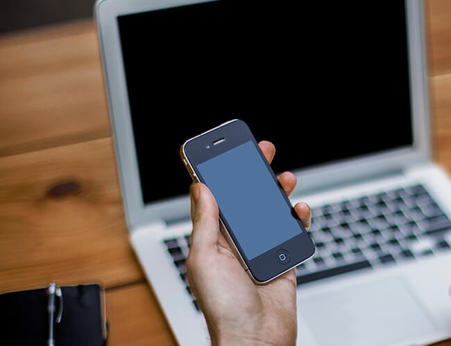 Hand holding a smartphone with a blank screen in front of an open laptop on a wooden desk.
