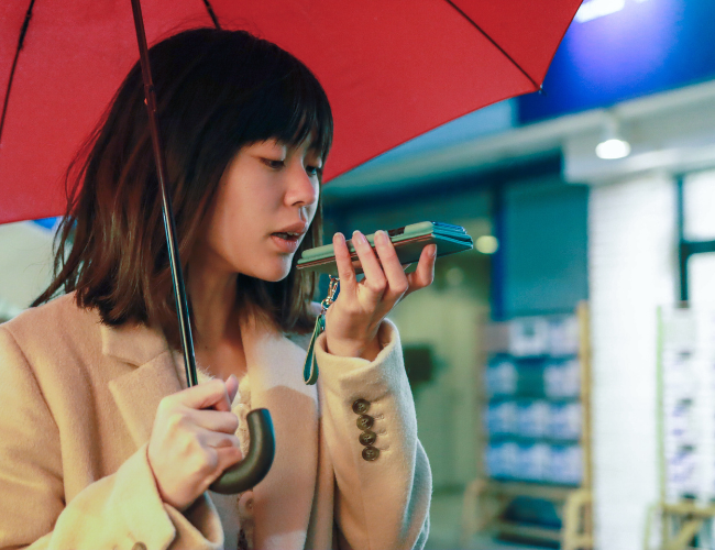 Woman holding a red umbrella speaking into a smartphone held close to her mouth on a city street.