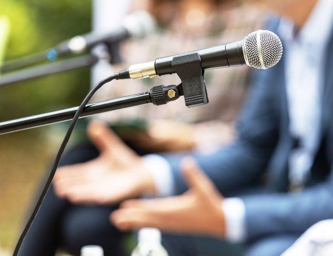 Close-up of a black handheld microphone on a stand with a blurred person gesturing while speaking in the background.