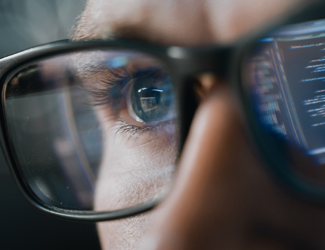 Extreme close-up of a person's eye behind glasses with lines of code reflected in the lenses.