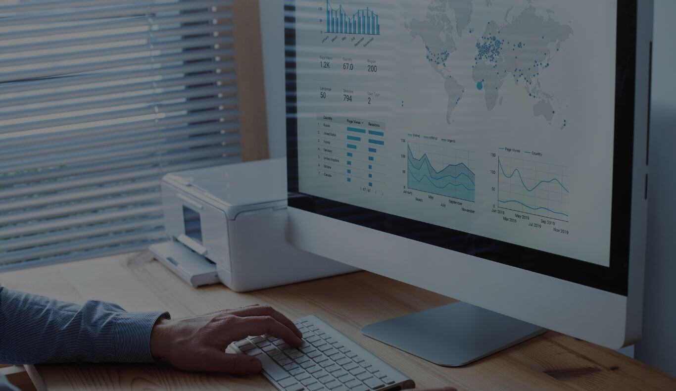Hand typing on a keyboard in front of a monitor displaying analytics dashboards with a world map and charts.