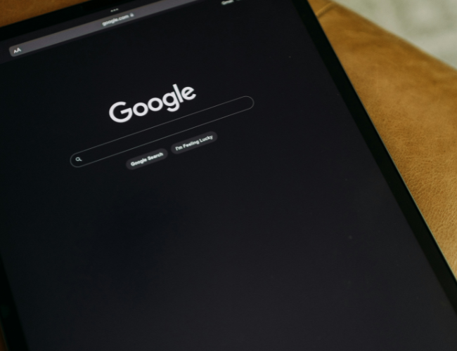 Tablet on a brown surface displaying the Google search homepage in dark mode.