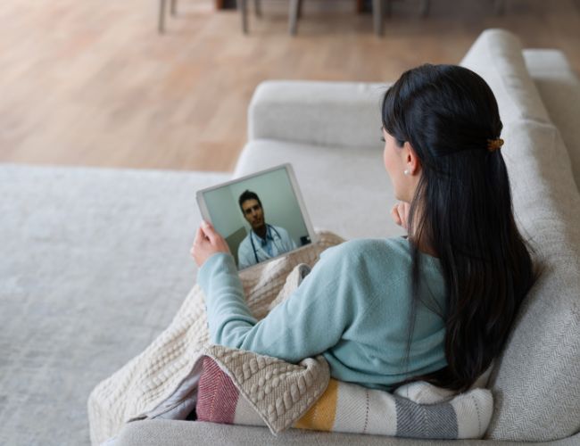 Woman in a teal sweater on a sofa holding a tablet displaying a video call with a male doctor wearing a stethoscope.