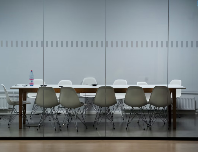 Empty modern conference room with a long wooden table surrounded by white and gray shell chairs.