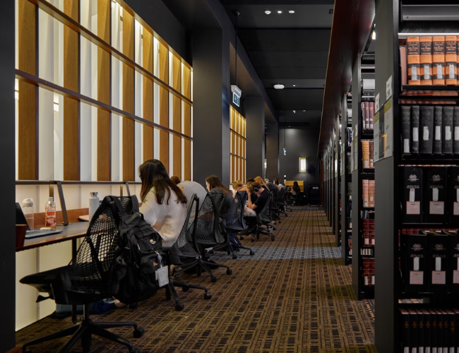 Students study at a row of carrels along a wall in a modern law library, with tall shelves of bound legal volumes lining the opposite side.