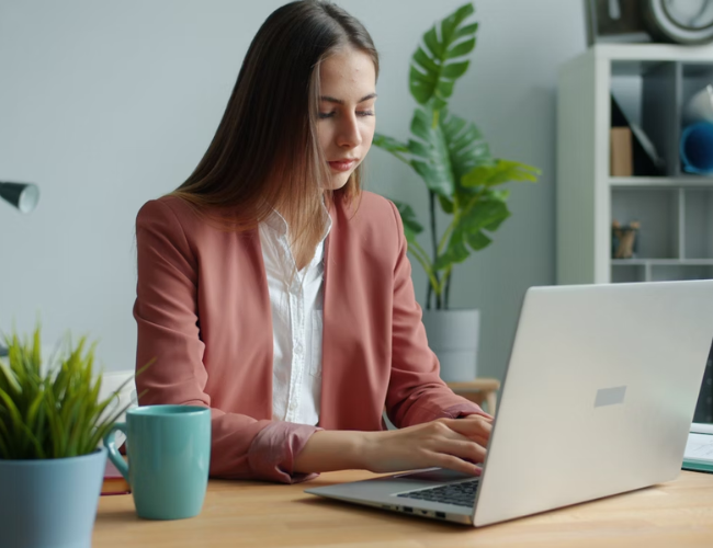 Young woman in a pink blazer typing on a laptop at a tidy desk with a green plant and blue coffee mug.